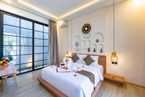 Romantic Bali villa bedroom with king platform bed decorated in rose petals and swan towel art, large glass doors to garden, wooden accents, and warm pendant lighting.